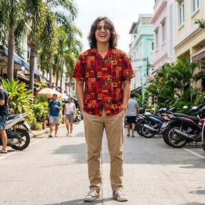 May include: A person wearing a short-sleeved, button-down shirt with a red background and a repeating pattern of orange and black tribal designs. The person is also wearing khaki pants and gray sneakers. The background shows a street with palm trees and parked motorcycles.