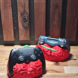May include: Two game controllers, one black and one blue, each resting on a red brain-shaped stand. The black controller has a green loop. The background is a wooden wall.