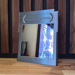 May include: A silver-colored wall mirror shaped like a classic Nintendo Game Boy game cartridge. The frame features the embossed text Nintendo GAME BOY at the top and a small triangle detail at the bottom center of the reflective glass surface.
