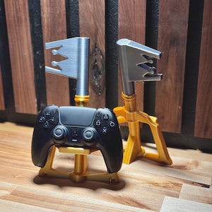 May include: A black video game controller rests on a yellow stand. The stand has a unique design with a silver emblem on top. Another stand is visible in the background. The stands are on a wooden surface.