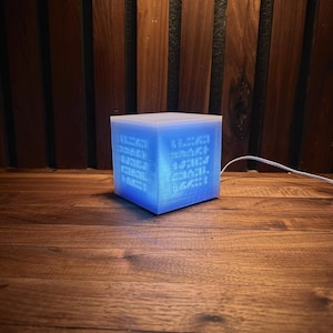 May include: A glowing blue cube-shaped lamp with etched patterns on its sides. The lamp emits a soft, ambient light, creating a calming atmosphere. The lamp is placed on a wooden surface, and a white cord extends from the back.