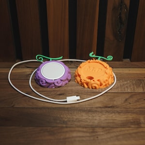 May include: Two colorful, fruit-shaped charging pads with green stems and a white charging cable. One pad is purple with a silver center, and the other is orange with the letters "M.P." on it. The pads are on a wooden surface.