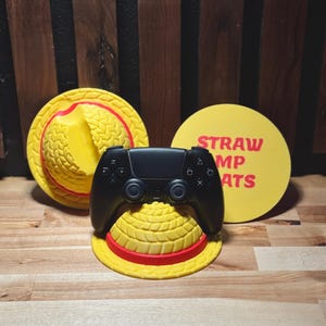 May include: A black game controller rests on a yellow straw hat with a red band. A yellow disc with the words "STRAW MP ATS" in red is behind the controller. Another yellow straw hat is visible in the background.