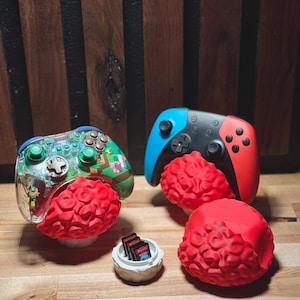 May include: Two game controllers, one blue and red, the other clear with green buttons, rest on red, brain-shaped stands. A small white container holds game cartridges. The controllers are for a gaming console.