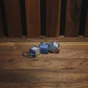 May include: A set of three blue cube-shaped keychains with a silver keyring. The keychains are on a wooden surface with a dark wooden background. The keychains have a textured appearance and a small handle.