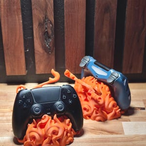 May include: Two game controller stands. One stand holds a black controller, the other holds a blue controller. Both controllers rest on orange flame-shaped bases. The background is a wooden wall.