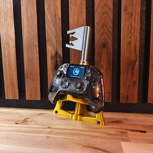 May include: A black transparent game controller with a central digital screen displaying a fox logo, mounted on a gold-colored plastic stand. A silver metal key-shaped attachment extends vertically from the top of the controller housing.