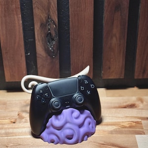 May include: A black video game controller rests on a purple brain-shaped base. The controller has buttons and directional controls. The base is a detailed, textured sculpture. The background is a wooden surface.