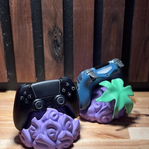 May include: Two game controller stands. One stand holds a black controller, the other a blue controller. Both controllers rest on a purple, textured base. The blue controller stand has a green leaf accent. The background is a wooden wall.