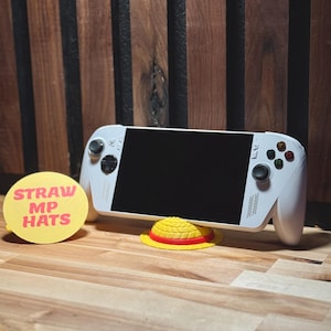 May include: A white handheld gaming console with a black screen, resting on a wooden surface. A yellow and red straw hat and a yellow circle with the text "STRAW MP HATS" are also present. The background is a wooden wall.