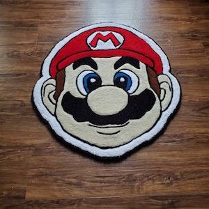 May include: A plush rug featuring a cartoon character with a red hat, black moustache, and blue eyes. The character is smiling and has a white outline.