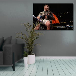 Könnte beinhalten: Gerahmter Leinwanddruck mit einem siegreichen Athleten in einem Boxring, der mit Meisterschaftsgürteln und einem roten Umhang geschmückt ist. Das Bild ist vor einem dunklen Hintergrund platziert, wobei der Triumphausdruck des Athleten deutlich sichtbar ist. Der Druck ist an einer grauen Wand befestigt.