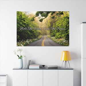 Op de afbeelding: Een canvas print met een schilderachtige weg die door een weelderig groen bos slingert. De afbeelding bevat de tekst "Hold The Vision, Trust The Process". De weg wordt geflankeerd door levendig groen en wilde bloemen.