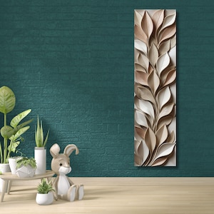 Könnte beinhalten: Ein vertikales Wandkunstwerk mit einem Reliefdesign aus stilisierten Blättern in Beige-, Creme- und Brauntönen. Das Kunstwerk ist auf einem weißen Hintergrund montiert und erzeugt einen dreidimensionalen Effekt. Das Stück wird vor einer tealfarbenen Backsteinmauer ausgestellt.