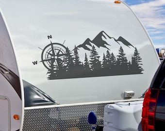 May include: A white camper van with a black and gray mountain and forest silhouette decal on the side. The decal includes a compass with the letters N, W, and E.