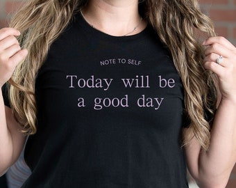 May include: Black crew neck t-shirt with the text "NOTE TO SELF Today will be a good day" in a light purple font. The shirt is worn with blue jeans. The image is of a person with long, wavy hair.
