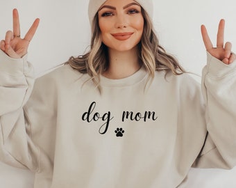 Könnte beinhalten: Beiges Sweatshirt mit dem Schriftzug "dog mom" in Schreibschrift und einem Pfotenabdruck. Das Sweatshirt hat eine neutrale Farbe und eine lockere Passform. Eine Mütze ist ebenfalls zu sehen.