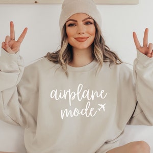 May include: Beige sweatshirt with the words "airplane mode" in white script and a small airplane graphic. The sweatshirt is a neutral color and has long sleeves. A person is wearing the sweatshirt and a beige beanie.