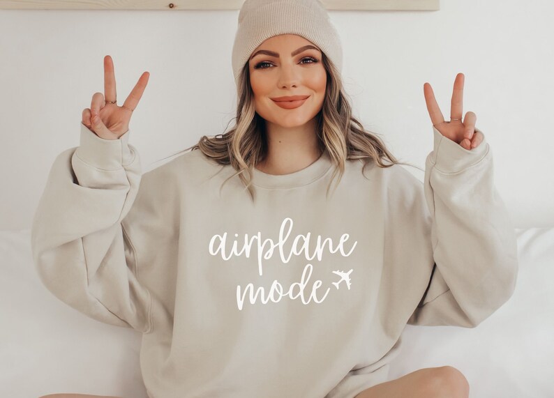 May include: Beige sweatshirt with the words "airplane mode" in white script and a small airplane graphic. The sweatshirt is a neutral color and has long sleeves. A person is wearing the sweatshirt and a beige beanie.