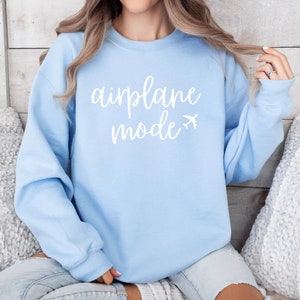 May include: A light blue sweatshirt with the words "airplane mode" in white script, accompanied by a small airplane graphic. The sweatshirt is a solid color and has long sleeves. The image is of a person wearing the sweatshirt.