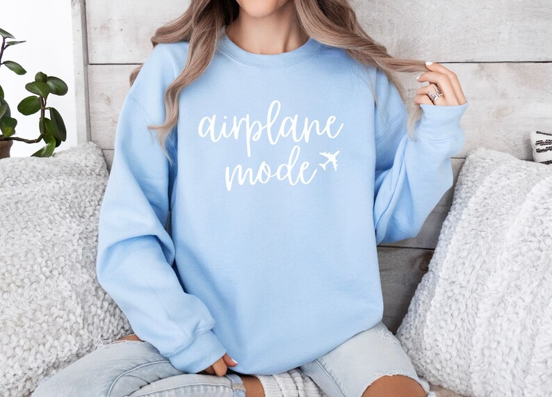 May include: A light blue sweatshirt with the words "airplane mode" in white script, accompanied by a small airplane graphic. The sweatshirt is a solid color and has long sleeves. The image is of a person wearing the sweatshirt.