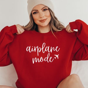 May include: A red sweatshirt with the words "airplane mode" in white script and a small airplane graphic. The long-sleeved top is a solid color and has a crew neck.