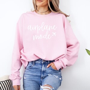 May include: A light pink sweatshirt with the words "airplane mode" in white script, along with a small airplane graphic. The long-sleeved top is paired with blue jeans.