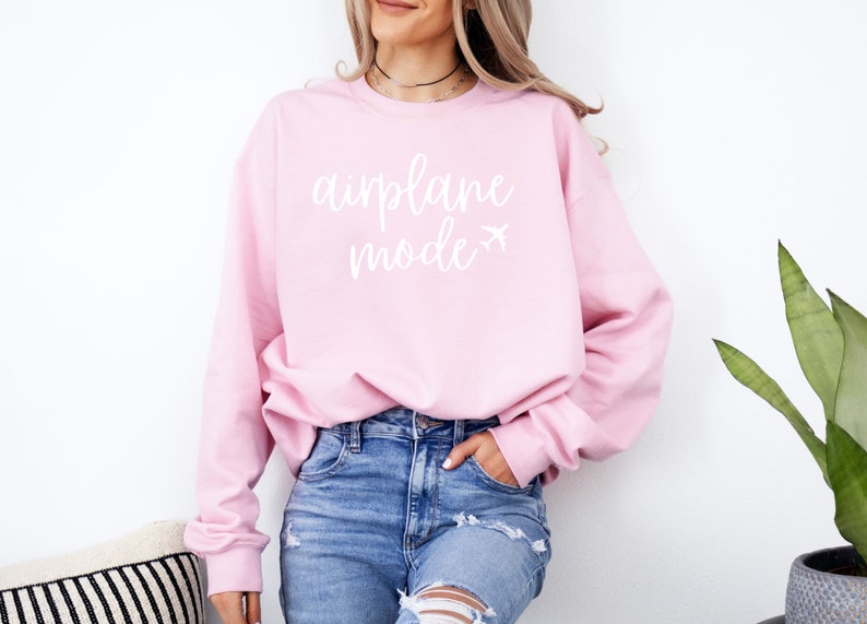 May include: A light pink sweatshirt with the words "airplane mode" in white script, along with a small airplane graphic. The long-sleeved top is paired with blue jeans.