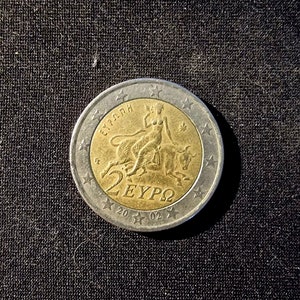 Op de afbeelding: Een zilveren en gouden twee euro munt met de afbeelding van een vrouw die op een stier rijdt. De munt heeft de tekst "ΕΥΡΩΠΗ" en "2 ΕΥΡΩ" erop.