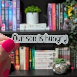 May include: A white sticker with the text "Our son is hungry" in a black, simple font. The sticker is held in front of a bookshelf filled with books and small plants.