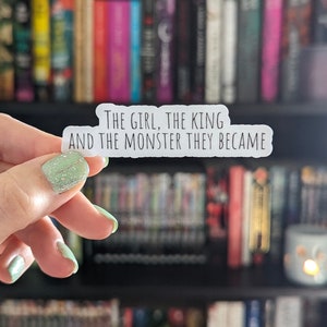 May include: A white sticker with the text "THE GIRL, THE KING AND THE MONSTER THEY BECAME" is held in front of a bookshelf filled with books. The sticker has a rounded rectangular shape and is held by a hand with green glitter nail polish.