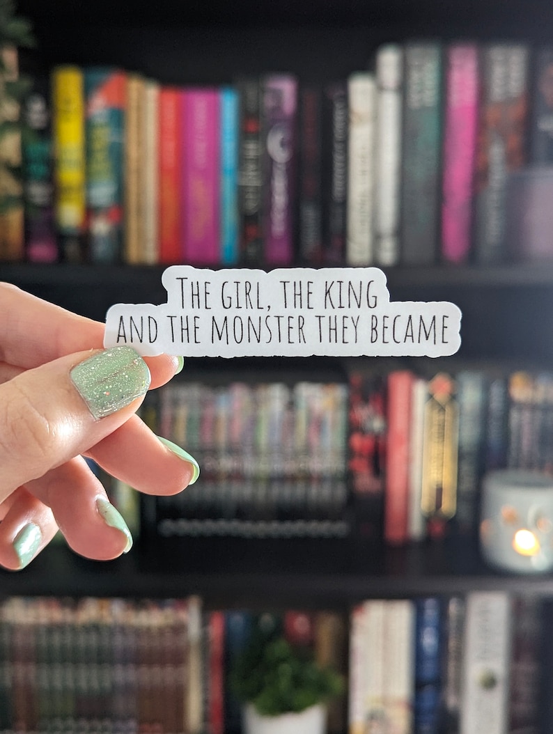 May include: A white sticker with the text "THE GIRL, THE KING AND THE MONSTER THEY BECAME" is held in front of a bookshelf filled with books. The sticker has a rounded rectangular shape and is held by a hand with green glitter nail polish.