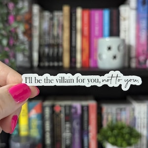 May include: A white sticker with the text "I'll be the villain for you, not to you" in black script. The sticker is held in front of a bookshelf filled with books of various colors. The person holding the sticker has pink nail polish.