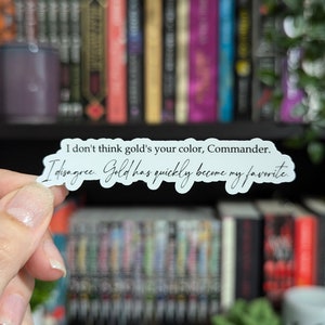 May include: A white sticker with black text that reads, "I don't think gold's your color, Commander. I disagree. Gold has quickly become my favorite." The sticker is held in front of a bookshelf.