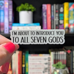 Puede incluir: Una pegatina blanca con texto negro que dice "I'M ABOUT TO INTRODUCE YOU TO ALL SEVEN GODS." La pegatina se sostiene frente a una estanter&iacute;a llena de libros y peque&ntilde;as plantas.