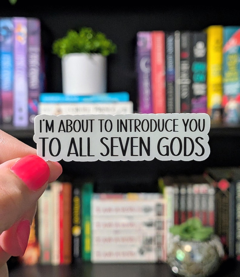 Puede incluir: Una pegatina blanca con texto negro que dice "I'M ABOUT TO INTRODUCE YOU TO ALL SEVEN GODS." La pegatina se sostiene frente a una estanter&iacute;a llena de libros y peque&ntilde;as plantas.