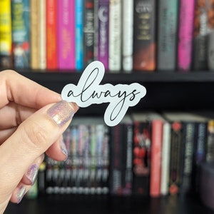 May include: A white sticker with the word "always" in a cursive font. The sticker is held in front of a bookshelf filled with books of various colors and sizes.