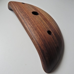 Puede incluir: Tabla de servir de madera en forma de media luna, con un acabado marrón oscuro. Tiene tres agujeros circulares. La veta de la madera es visible, añadiendo textura a la superficie lisa y curvada. La tabla está diseñada con un asa en un extremo.