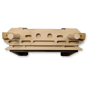 May include: A wooden climbing hold with multiple grip options. The hold is rectangular with various holes and rounded grips. Two cylindrical wooden dowels are attached on each end. The hold is light brown with dark brown accents.