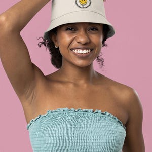 Puede incluir: Un sombrero de pescador beige con el texto "Under & Overstimulated" y una carita sonriente amarilla. La persona lleva un top de tubo azul claro. El fondo es rosa.