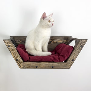 May include: A white cat sits on a wall-mounted cat bed. The bed is made of dark wood with a burgundy cushion. The bed has a unique geometric shape and is attached to a white wall.