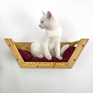 May include: A white cat sits in a wooden cat bed with a red cushion. The bed is mounted to a white wall.