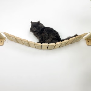 May include: A wooden cat bridge with a light-colored finish, suspended between two wall-mounted platforms. The bridge is made of small wooden planks and rope. A fluffy gray cat is resting on the bridge.
