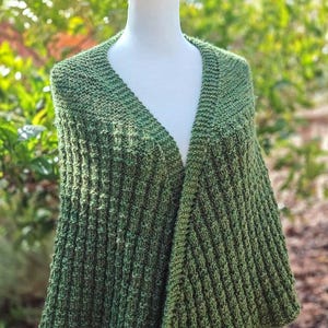 May include: A green knitted shawl displayed on a mannequin. The shawl has a textured, ribbed pattern and a V-neck design. The color is a mix of green shades, and the shawl is draped over the mannequin's shoulders.