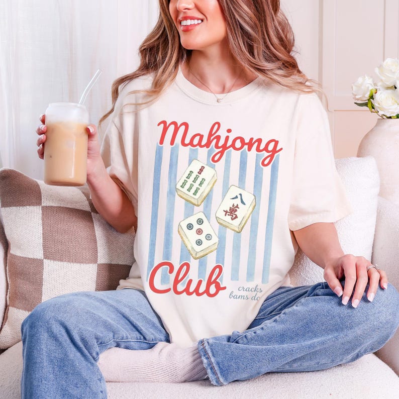 Puede incluir: Camiseta color crema con las palabras "Mahjong Club" en escritura roja. La camiseta presenta un gr&aacute;fico de tres fichas de Mahjong sobre un fondo de rayas verticales azules. Las palabras "cracks bams dots" est&aacute;n escritas en letra peque&ntilde;a.