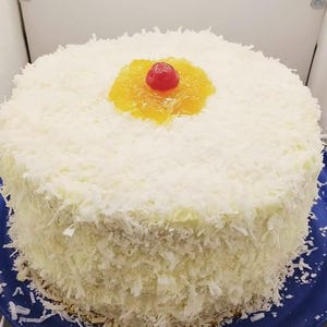 May include: A white cake covered in shredded coconut with a pineapple and cherry topping.