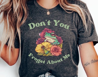 Puede incluir: Una camiseta gris oscuro con el texto "Don't You Froget About Me" en letras verdes. Una rana verde se sienta sobre una flor amarilla con flores rosas y rojas que la rodean.