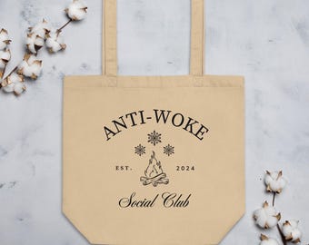 May include: A beige canvas tote bag with black text that reads "Anti-Woke Social Club" with a campfire and snowflakes graphic. The text also includes "Est. 2024".