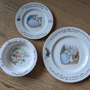 May include: A set of three white ceramic plates featuring illustrations from the children's book "The Tale of Peter Rabbit" by Beatrix Potter. The plates feature Peter Rabbit, Mrs. Rabbit, and Mr. McGregor. The plates are decorated with a blue and green border and have text from the book.