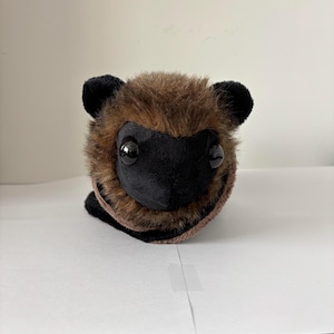 Puede incluir: Un peluche con forma de animal pequeño y redondo. El juguete tiene un cuerpo marrón y peludo, orejas negras y una cara negra con ojos grandes y redondos. El juguete está sobre una superficie blanca.